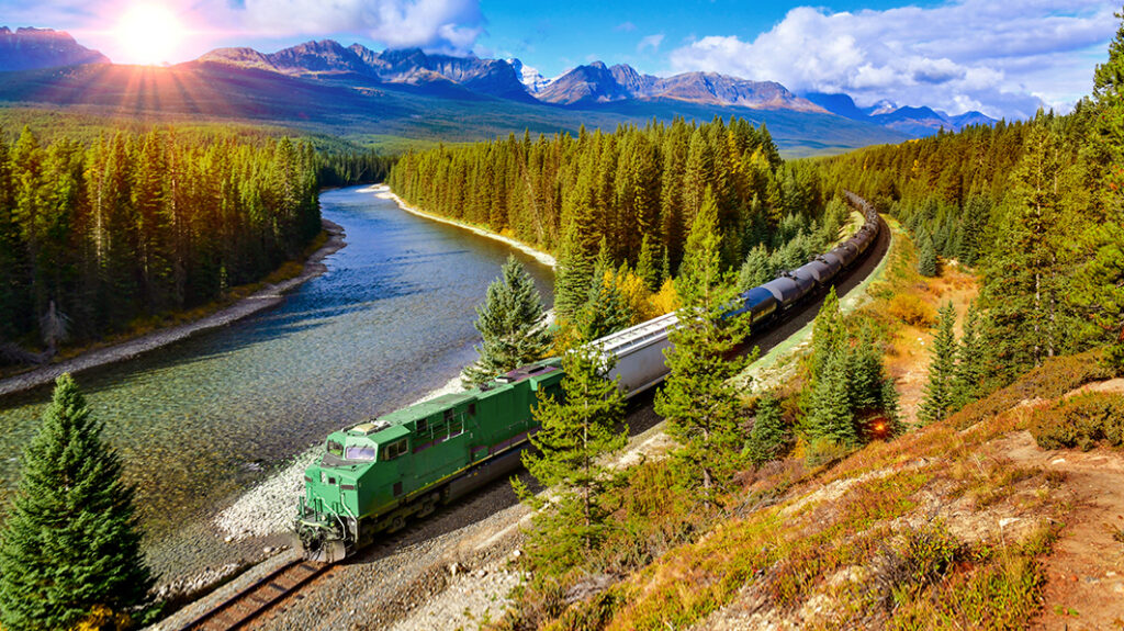 Scenic view of a green freight train winding through a forested mountain landscape beside a river, with the sun setting in the background